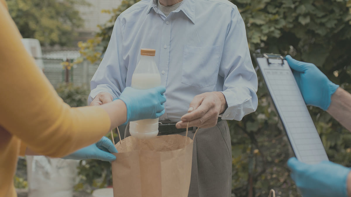 Volunteers in gloves handing a bag of groceries and milk to an older man