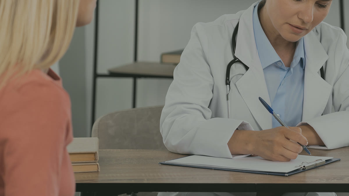 A doctor writing notes while talking with a patient across a desk